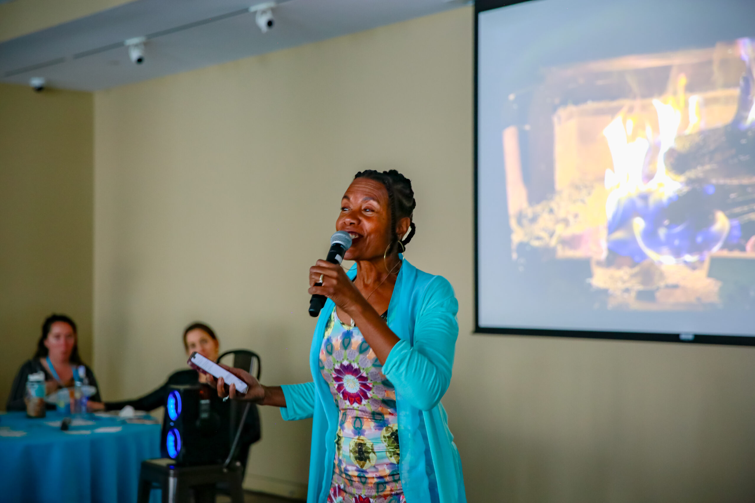 Leilani dressed in a Brain Jewells dress and open front turquoise long sleeve cover holds a microphone to her mouth. Behind Leilani is a screen showing a fire and a table with a few participants looking on.